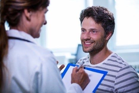 Um homem sorrindo enquanto conversa com uma profissional de saúde, que segura prancheta. A imagem sugere uma consulta médica que poderia incluir procedimentos como bichectomia para melhorar o contorno facial.