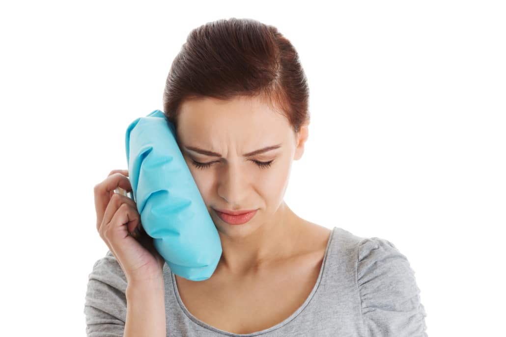 Mulher com expressão de desconforto, segurando uma bolsa de gelo na bochecha para aliviar a dor após a extração do siso, isolada contra um fundo branco.