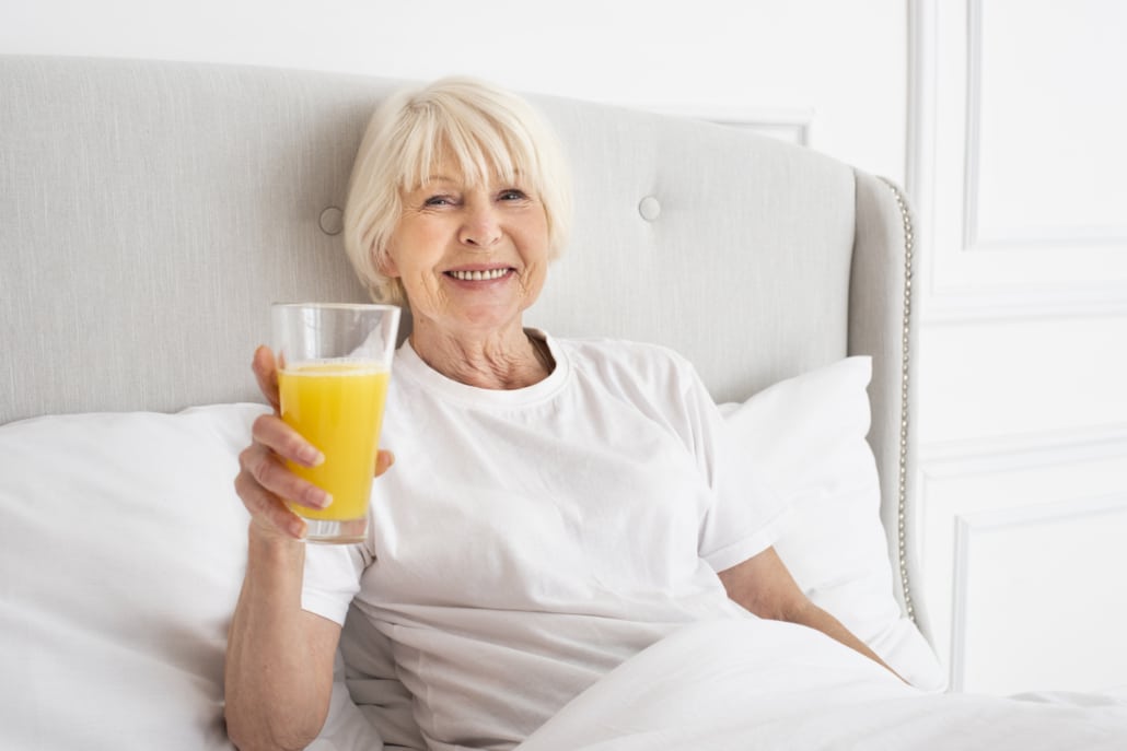 senhora idosa sorridente, segurando um copo de suco de laranja enquanto está sentada confortavelmente em sua cama, representando um estilo de vida saudável após a recuperação de um procedimento de enxerto ósseo.