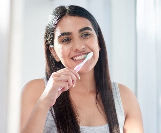 Jovem sorridente escovando os dentes em um ambiente bem iluminado. Seu sorriso radiante e dentes saudáveis sugerem uma rotina de cuidado dental que pode incluir a manutenção de implantes dentários com carga imediata.