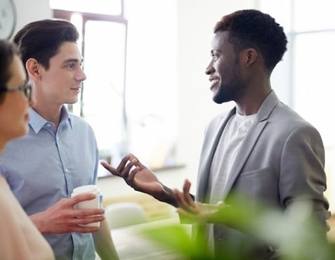 Mostra dois homens, um negro e um caucasiano, em um ambiente de escritório com janelas amplas ao fundo, envolvidos em uma conversa amigável. O homem negro, vestindo um terno cinza, está falando e gesticulando com uma mão, enquanto o caucasiano, de camisa azul, segura um copo de café e ouve atentamente.