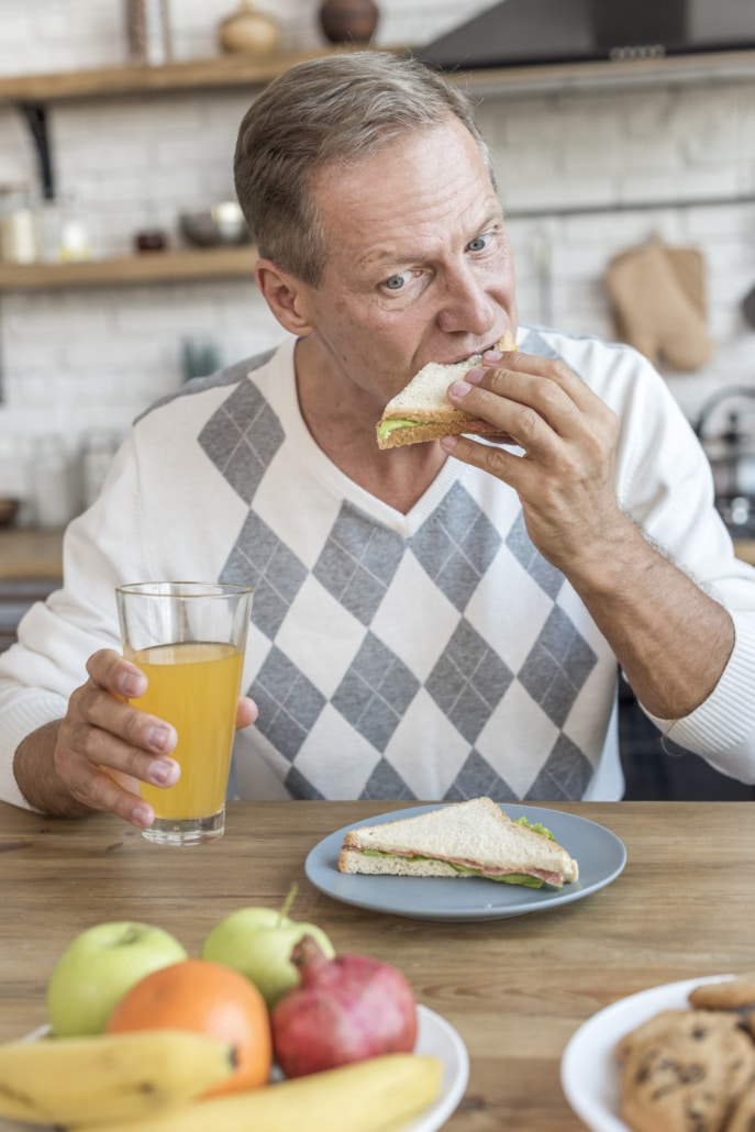 Um homem mais velho, com cabelos grisalhos e usando um suéter de padrão argyle, está sentado à mesa de uma cozinha moderna, comendo um sanduíche e bebendo suco de laranja. A mesa também tem uma variedade de frutas, sugerindo uma refeição saudável.