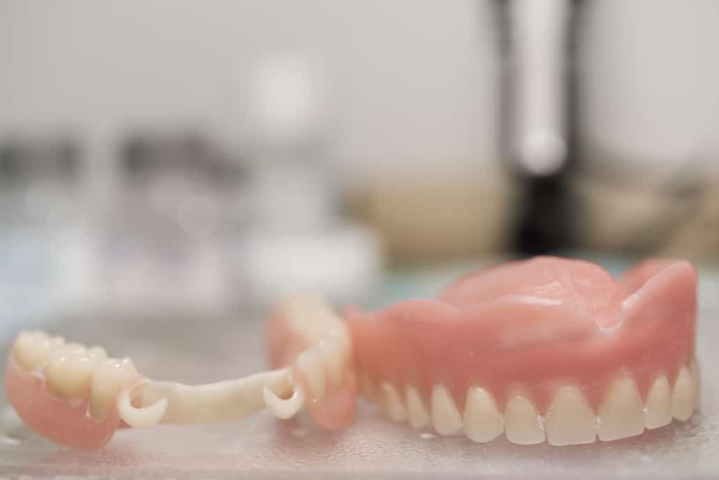 A imagem mostra uma prótese dentária fixa sobre uma bancada, possivelmente em um laboratório dental. A prótese, que simula a estrutura completa de uma mandíbula inferior, inclui dentes artificiais de cor natural fixados em uma base rosada que imita a gengiva. Este tipo de prótese é usado para substituir dentes perdidos, restaurando a função mastigatória e a estética do sorriso.