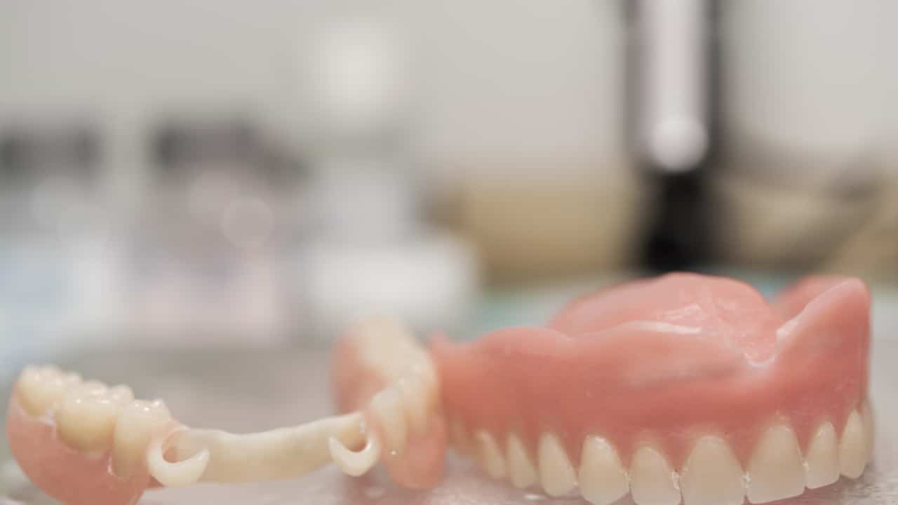 A imagem mostra uma prótese dentária fixa sobre uma bancada, possivelmente em um laboratório dental. A prótese, que simula a estrutura completa de uma mandíbula inferior, inclui dentes artificiais de cor natural fixados em uma base rosada que imita a gengiva. Este tipo de prótese é usado para substituir dentes perdidos, restaurando a função mastigatória e a estética do sorriso.