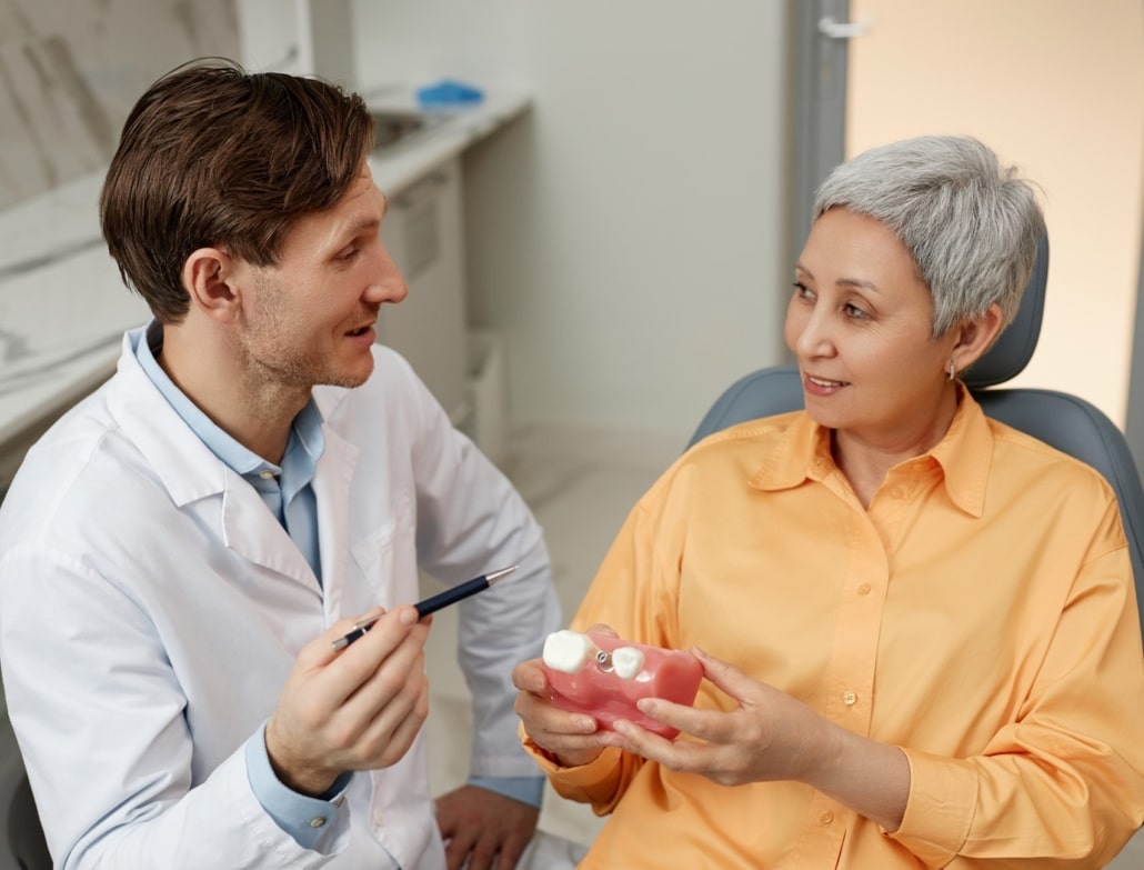  Um dentista orientando uma paciente sênior sobre a importância dos implantes dentários após anos de perda dentária. A paciente segura um modelo de prótese dentária, mostrando o processo de inserção do implante. 