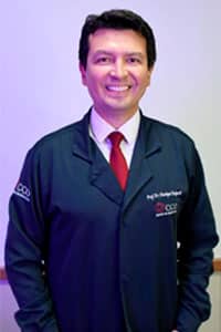 Dr. Henrique Taniguchi vestindo um jaleco azul olhando e sorrindo para a câmera