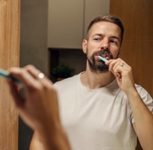 Um homem com barba, usando uma camiseta branca, escova os dentes em frente ao espelho do banheiro. Ele sorri sutilmente enquanto realiza sua rotina de higiene bucal, enfatizando a importância de cuidados diários para nunca mais ter cárie.