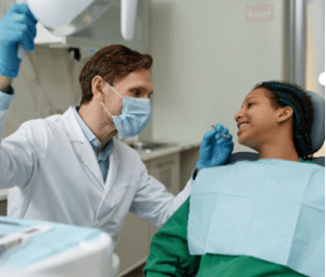  Um dentista de máscara conversando amigavelmente com uma paciente jovem na cadeira odontológica, demonstrando cuidado e atenção. Avaliar o siso é parte essencial das consultas para garantir a saúde oral completa dos pacientes. 